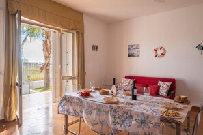 Un comedor con una mesa preparada para dos personas, un sofá rojo y vistas a una palmera a través de una puerta abierta.
