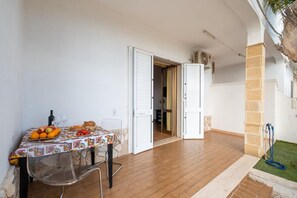 Comedor con mesa lista para dos, una silla y mantel. Se ve una puerta que da a otra habitación y un pequeño jardín con una manguera.