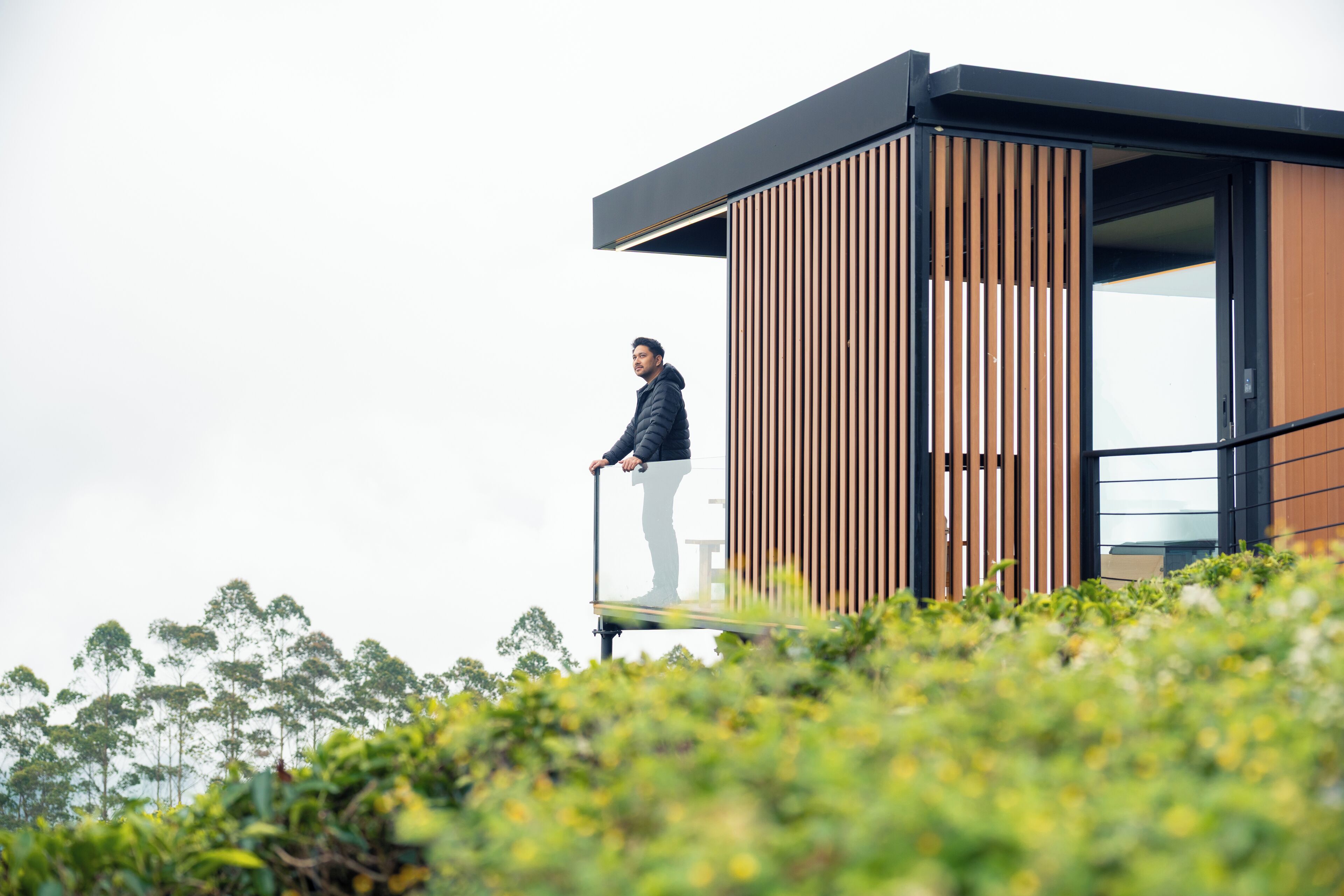 A person standing on a balcony of a modern building with vertical wooden slats and large windows.
