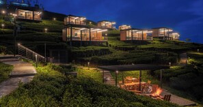 Une propriété située sur une colline, dotée de constructions modernes en verre, d’un foyer extérieur et d’un chemin éclairé.