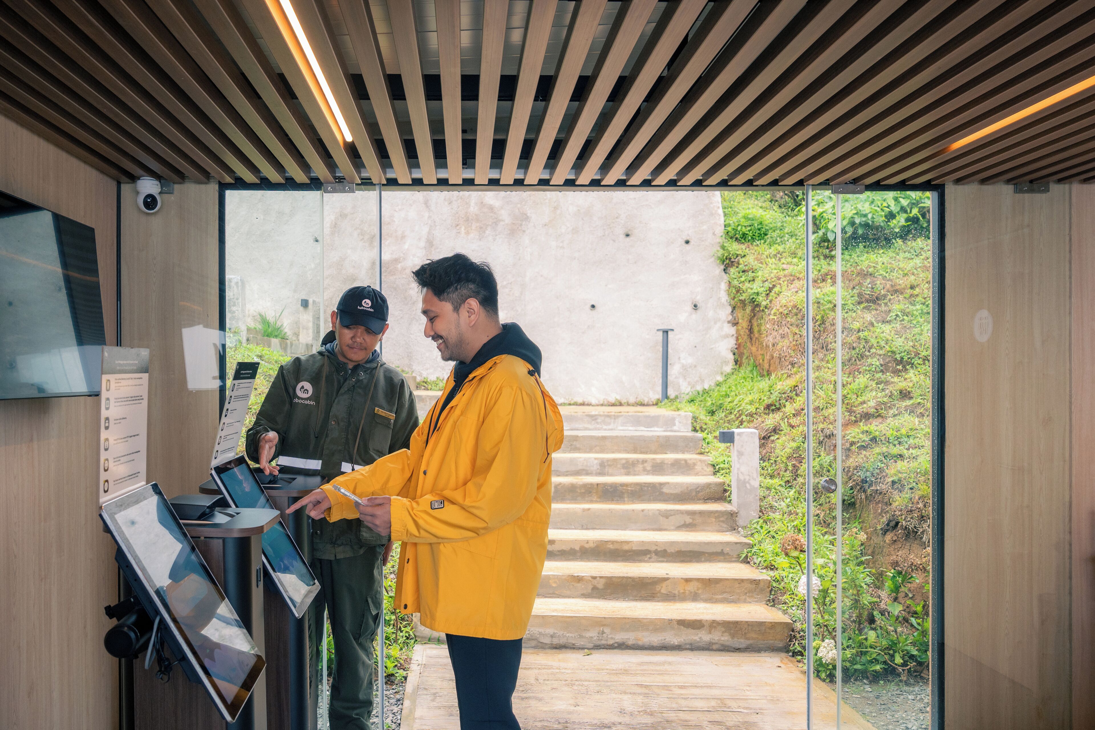 Deux personnes interagissent avec un kiosque numérique dans un intérieur moderne, avec vue sur un escalier extérieur et de la verdure.
