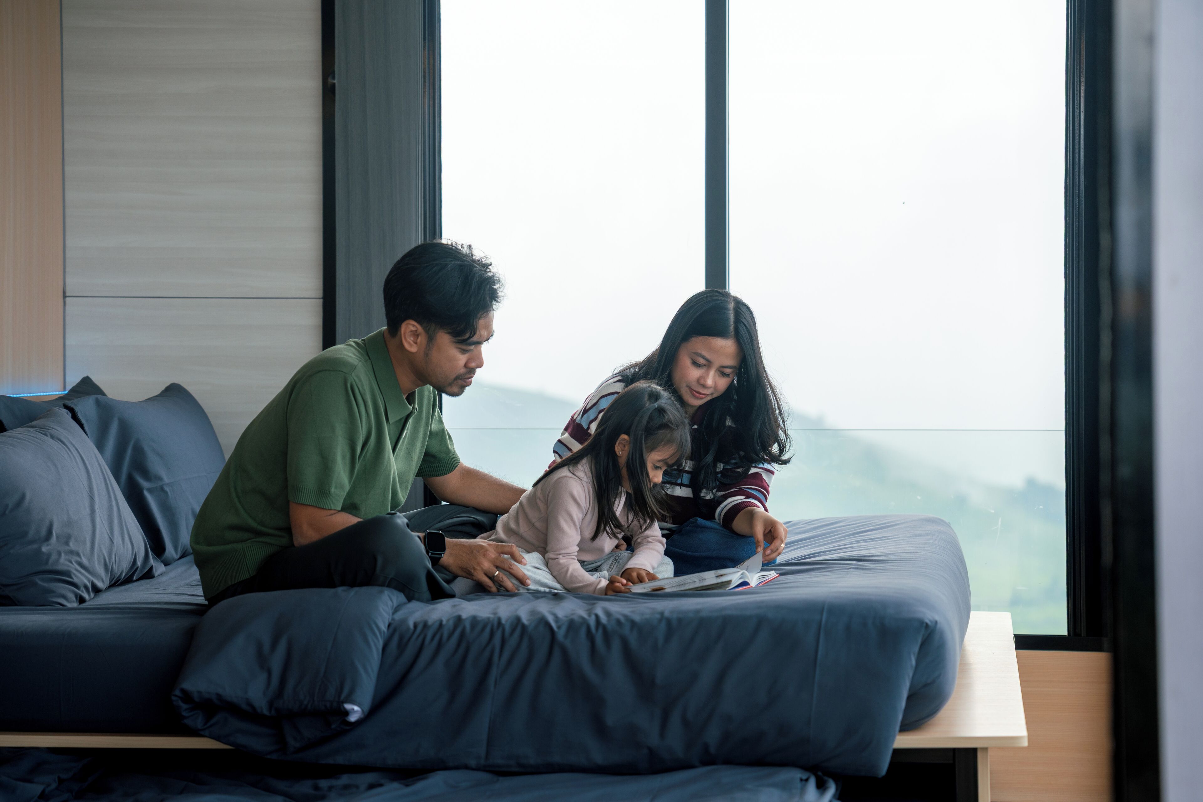 A family of three on a bed with a view of the outdoors.