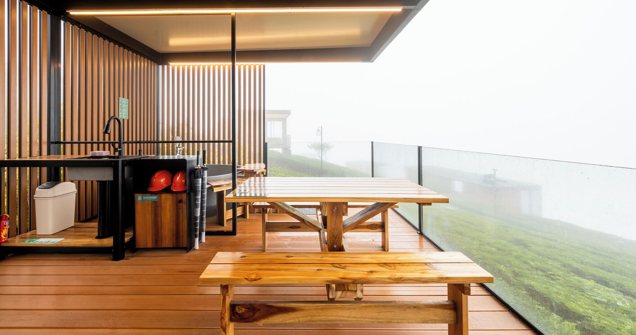 A modern outdoor dining area with wooden tables and benches, a small kitchen setup, and a view of a green landscape.