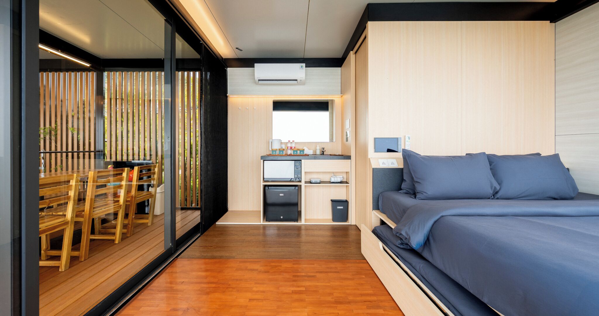 A modern bedroom with a bed, a small kitchenette, and a dining area with wooden furniture.
