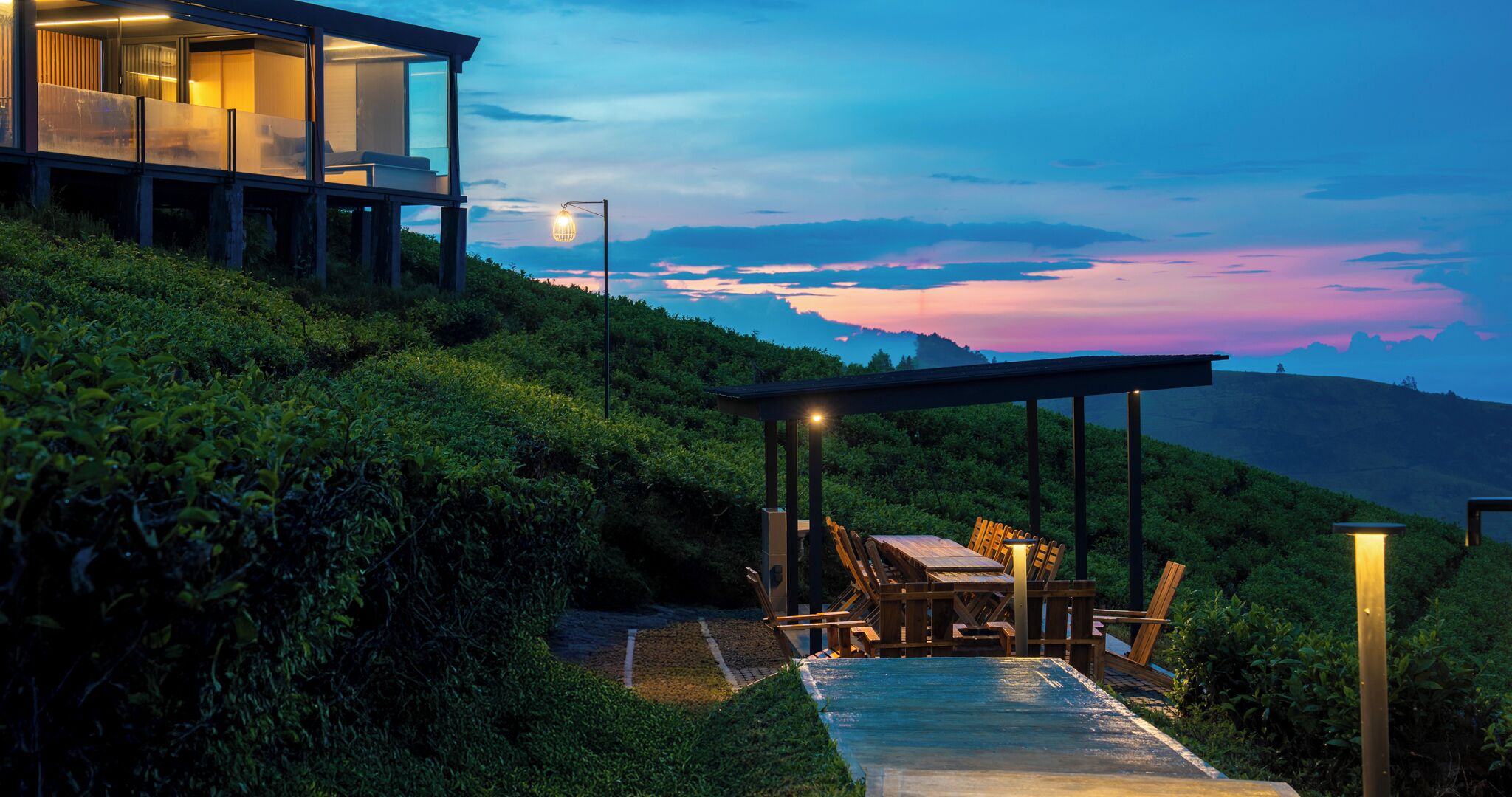 Uma casa moderna com fachada de vidro, área de jantar com mesa e cadeiras, e vista do pôr do sol sobre uma paisagem montanhosa.