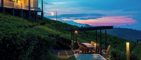 A modern house with a glass exterior, a dining area with a table and chairs, and a view of the sunset over a hilly landscape.