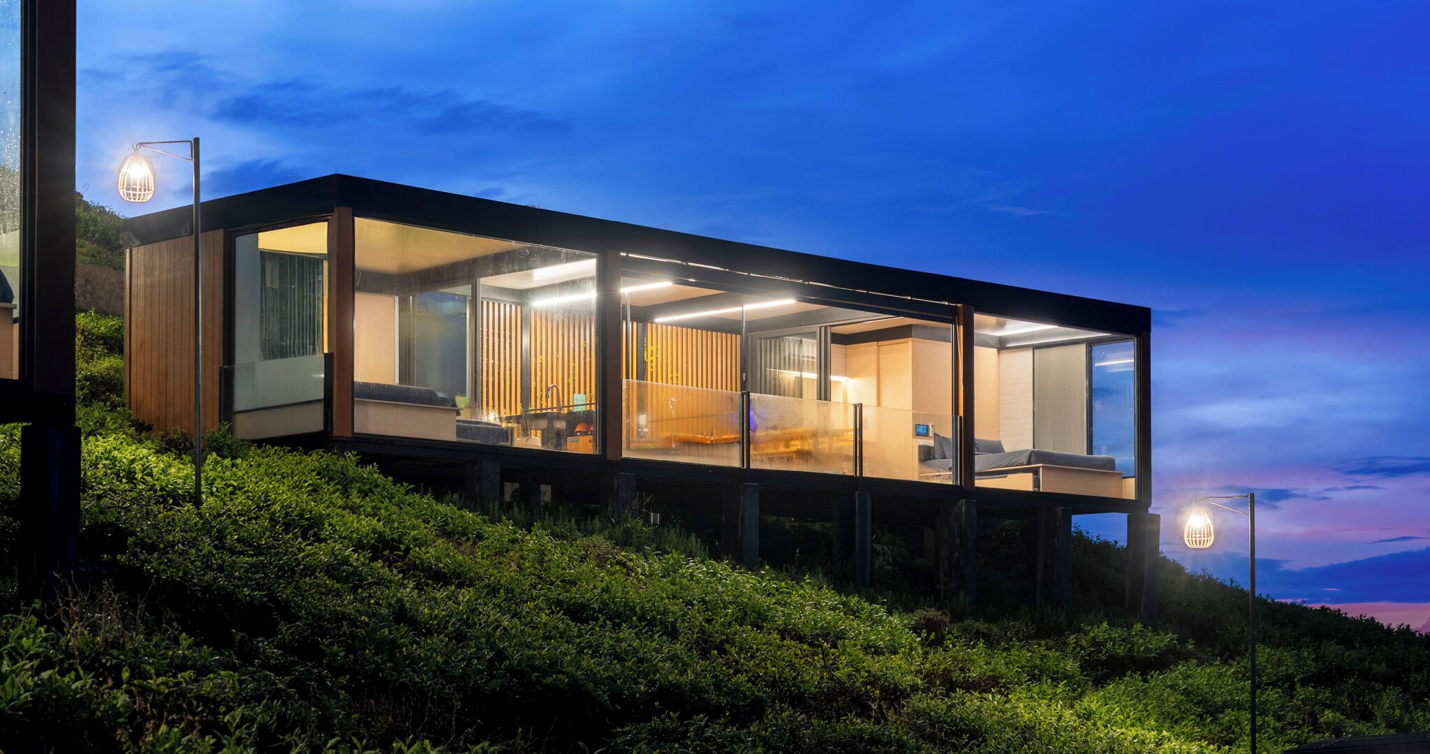A modern, glass-walled house with a visible kitchen and living area, situated on a hillside with lush greenery.