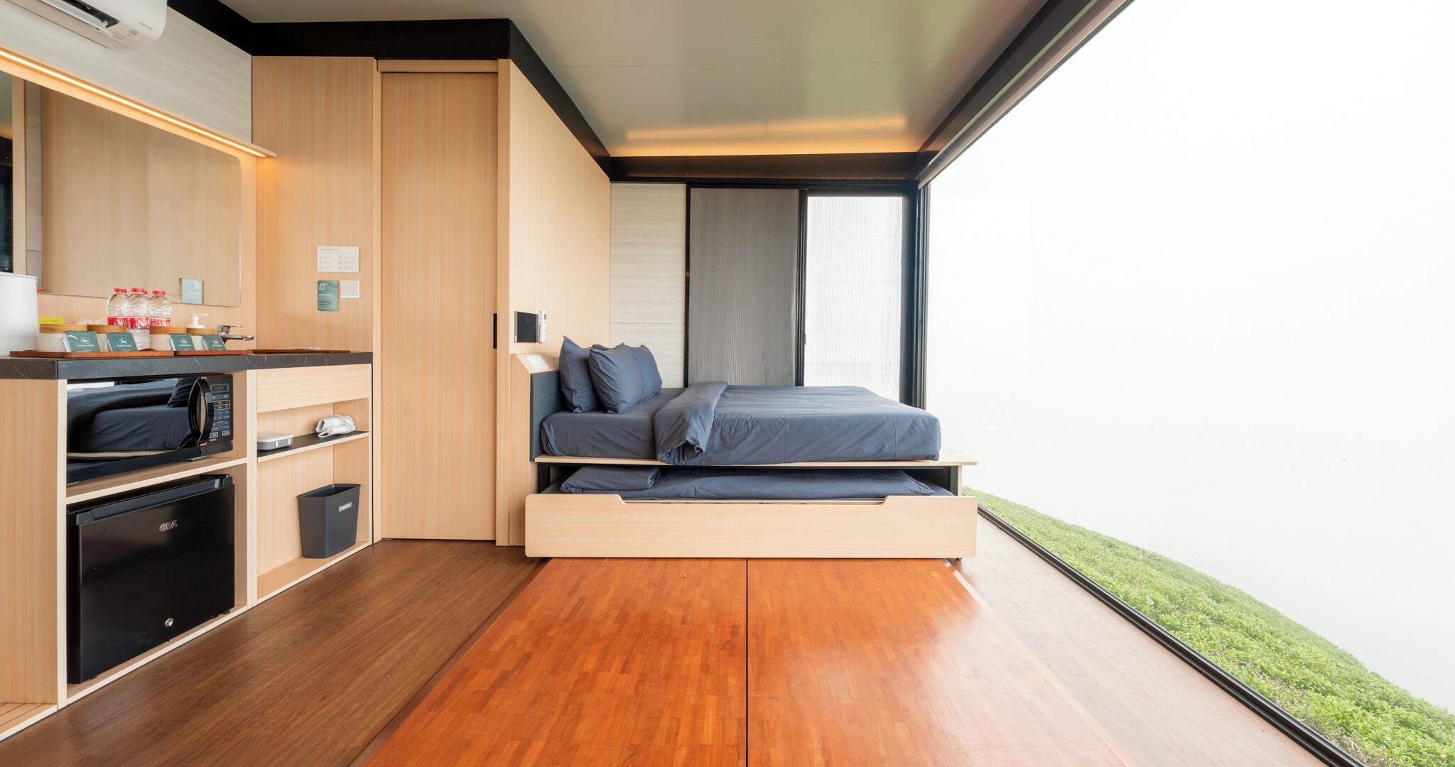 A compact room with a bed, kitchenette, and a view of greenery.