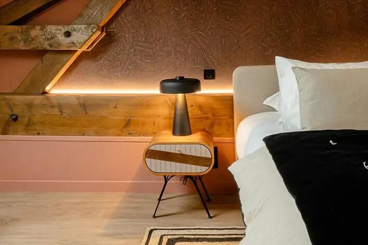 Quarto de sótão acolhedor com vigas de madeira aparentes, luminária moderna, cama com roupa de cama branca e tapete estampado.