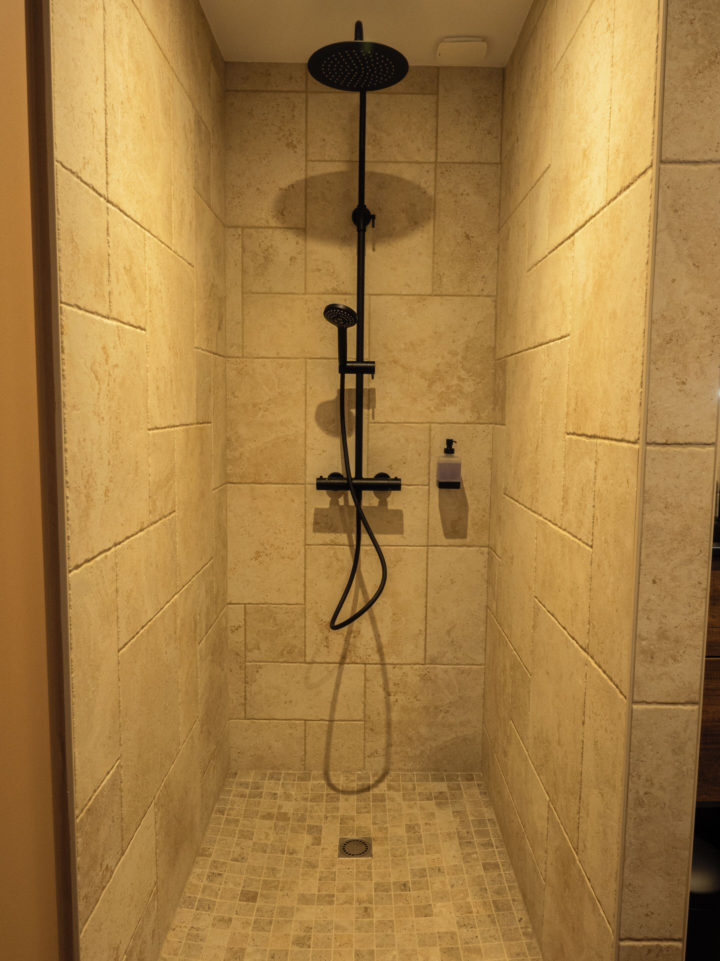 Eine kleine, mit Fliesen ausgekleidete Duschkabine mit Handbrause und am Wand montiertem Wasserhahn.