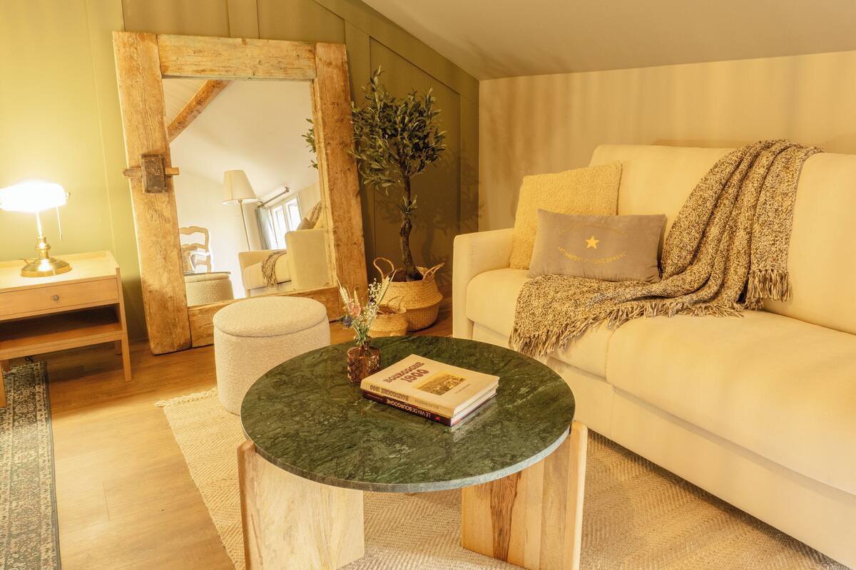 Uma sala de estar aconchegante com uma mesa de centro redonda de madeira, um sofá branco com um cobertor e um espelho com moldura de madeira.