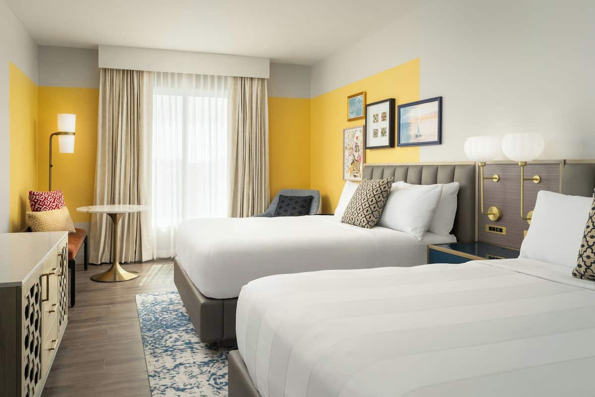 A hotel room with two beds, a small table, a lamp, and a view of the outdoors through a window.