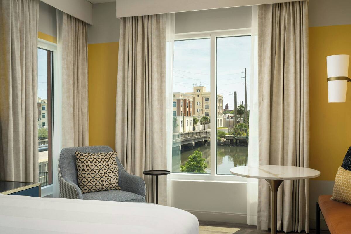 A hotel room with a large bed, a seating area with a table, and a view of a cityscape through a window.