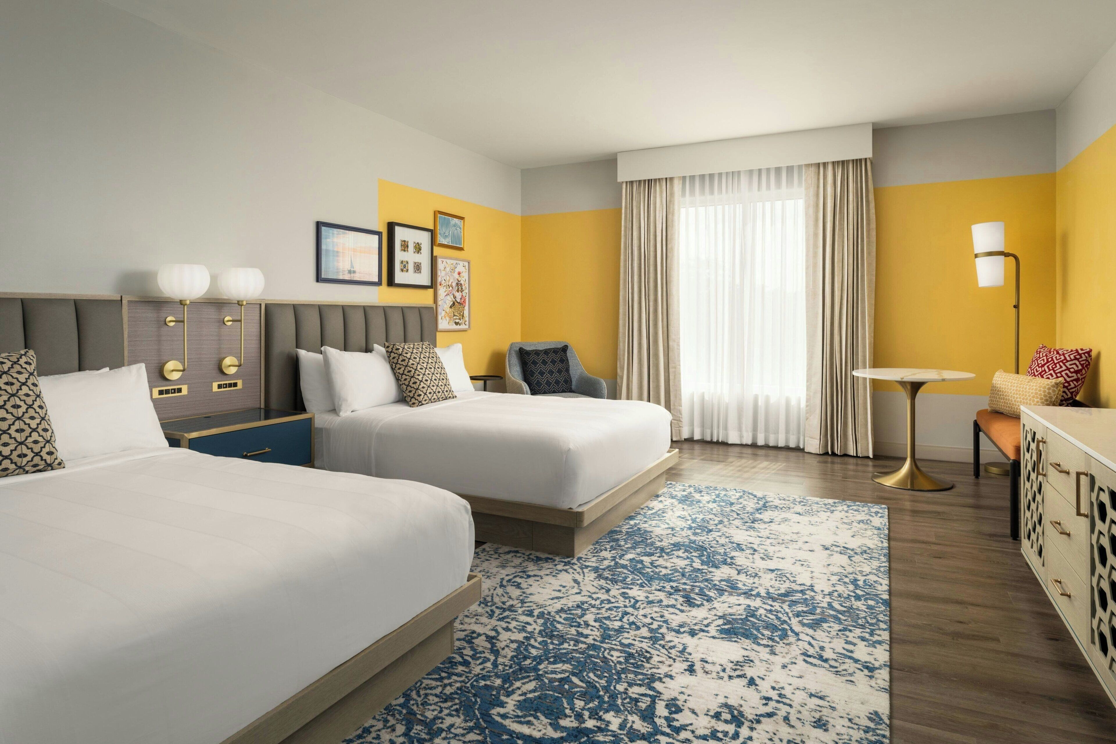 A hotel room with two beds, a desk, a chair, a small table, and a lamp.