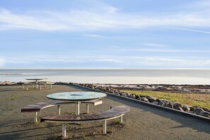 Ein Sitzbereich direkt am Strand mit Tischen und Bänken, ein Kiesweg und Blick aufs Meer.