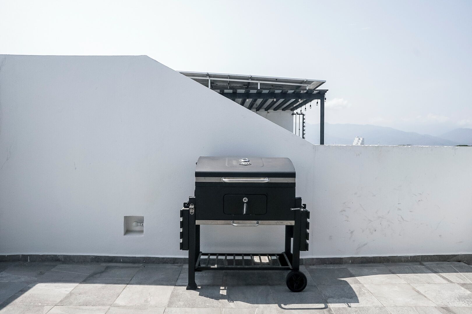 Una parrilla portátil en una terraza en la azotea, con una pared blanca y una pérgola de metal al fondo.