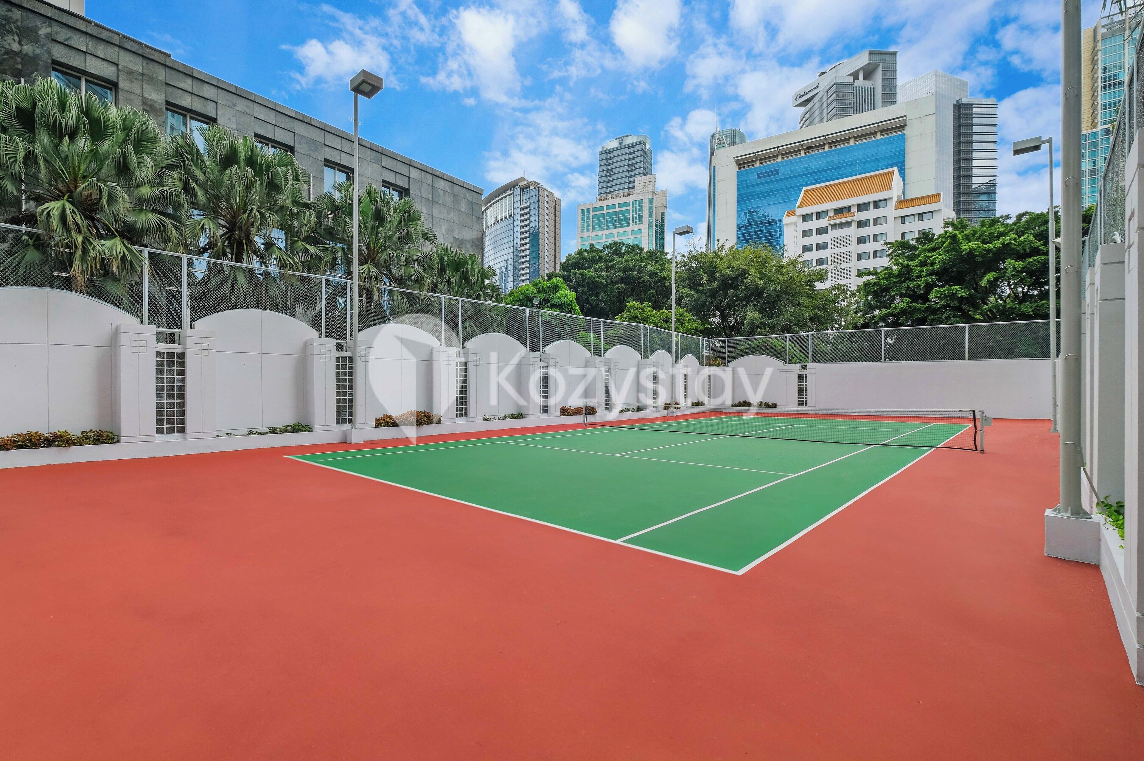 An indoor tennis court with a red and green surface, surrounded by a white fence and palm trees.