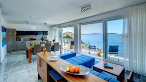 Un salón moderno con un ventanal grande con vistas al mar, un comedor con mesa de madera y una cocina al fondo.