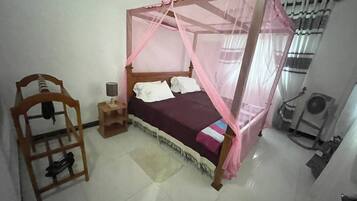 A bedroom with a bed under a pink canopy, a wooden chair, a small bedside table with a lamp, and a standing fan.