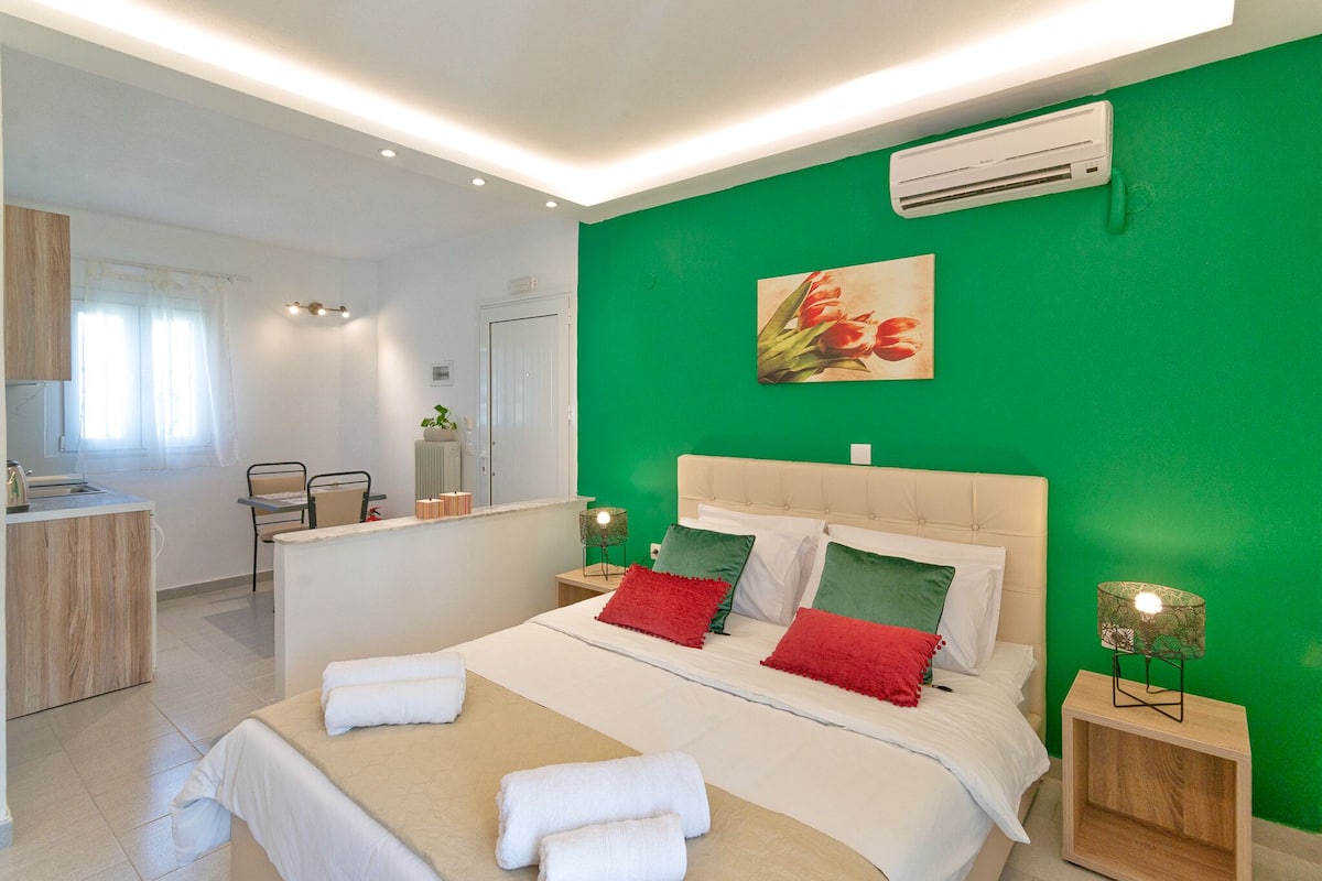 A modern bedroom with a green accent wall, a large bed with white and red bedding, a bedside table with a lamp, and a view of the adjacent bathroom.