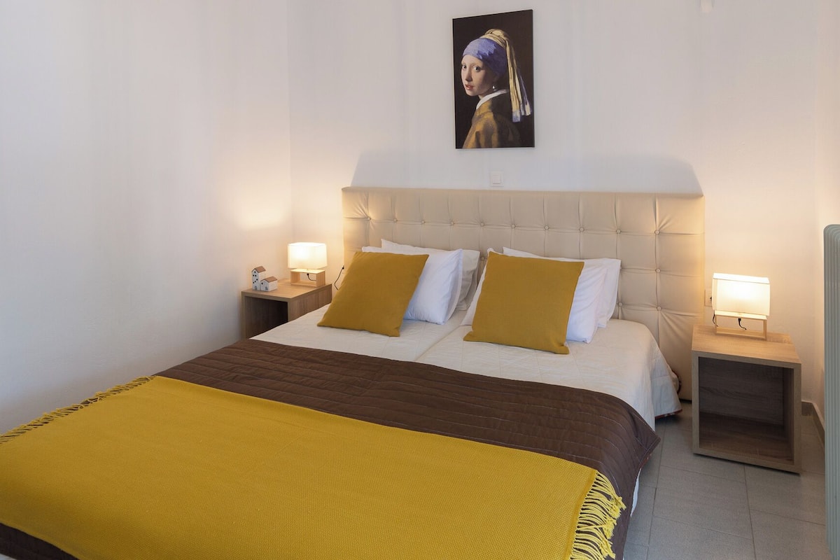 A neatly made bed with a brown quilt and a yellow throw blanket, two bedside lamps, and a painting above the headboard.
