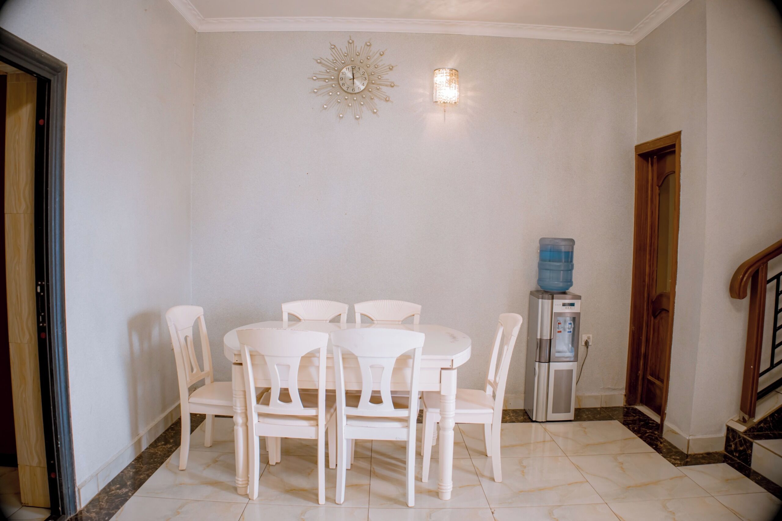 Uma área de refeições com mesa e cadeiras brancas, um dispensador de água e um relógio de parede.