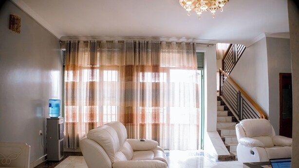 A spacious living room with a sofa, a chair, a water dispenser, and a staircase leading to an upper floor. - Wins Homes (Kigali)