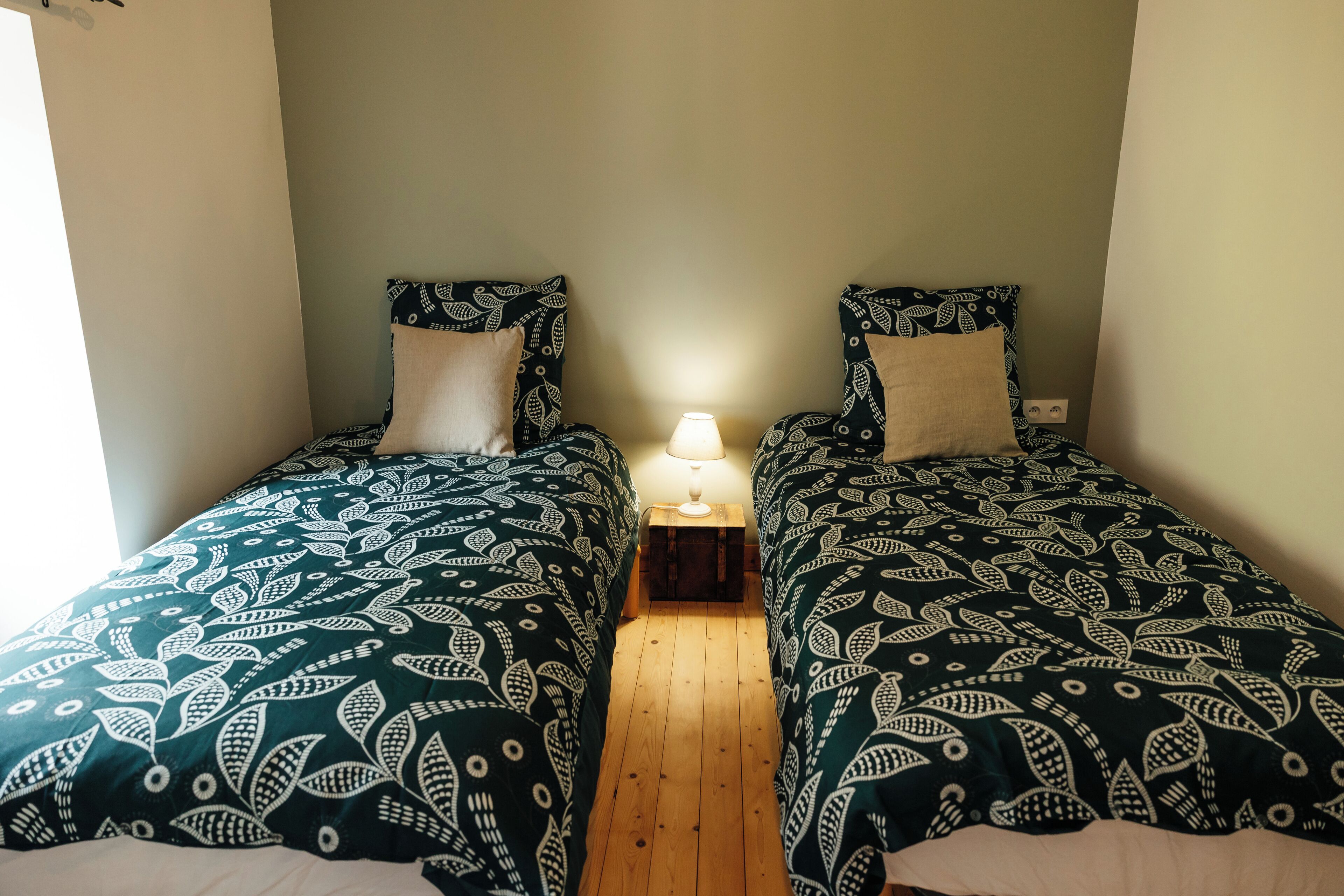 Una habitación con dos camas, cada una con ropa de cama estampada, una mesita de noche con lámpara y suelo de madera.
