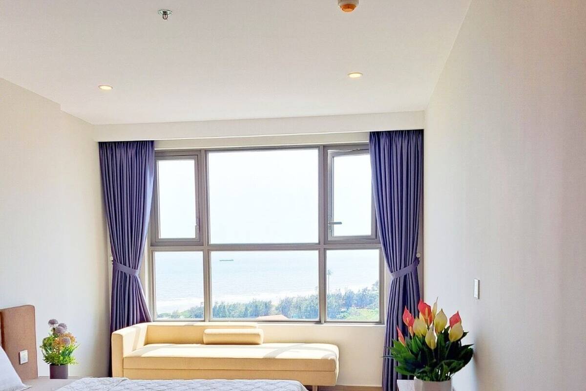 A bedroom with a bed, a sofa, a window with curtains, a bedside table, and a potted plant.