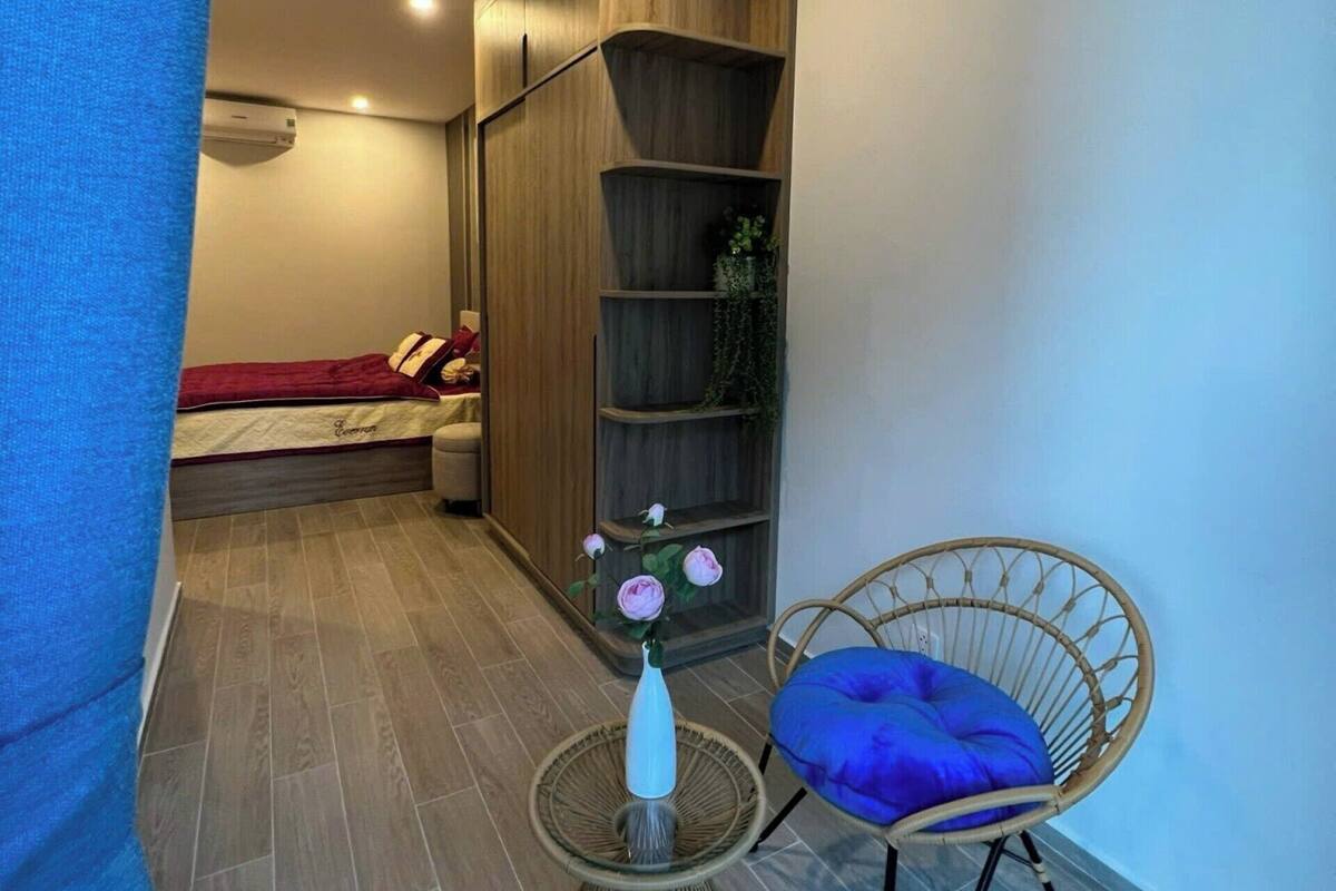 A compact room with a bed, a wooden shelving unit, a wicker chair with a blue cushion, and a small round table with a white vase.
