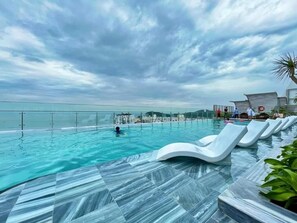 Ein Dachpool mit Infinity-Kante und freiem Blick auf das Meer, weiße Liegestühle und ein modernes Gebäude im Hintergrund.