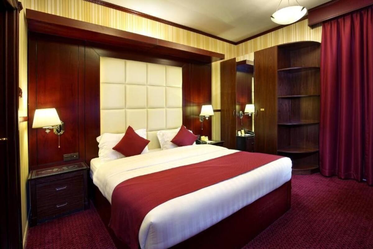 A large bed with a tufted headboard, two bedside tables with lamps, a red carpeted floor, and a wooden wardrobe with a mirror.