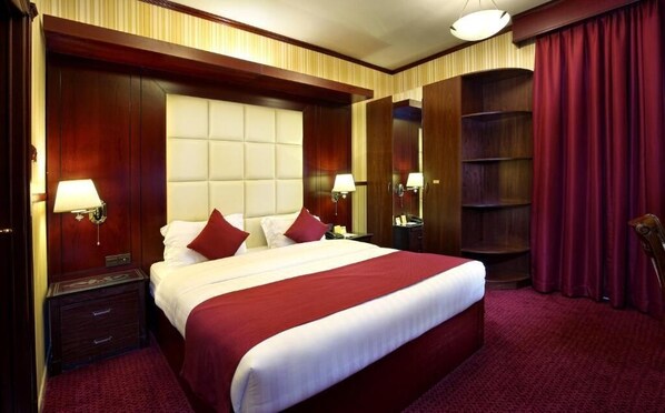 A large bed with a tufted headboard, two bedside tables with lamps, a red carpeted floor, and a wooden wardrobe with a mirror. - Benta Star Hotel LLC (Dubai)