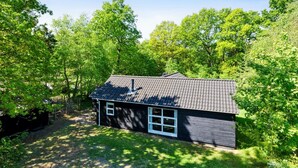 Een klein, donker huis met een pannendak, omgeven door weelderige groene bomen en gras.
