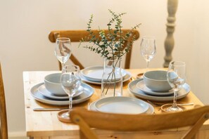 Una mesa de comedor de madera dispuesta con platos, tazones y copas de vino, junto a un jarrón con follaje verde.