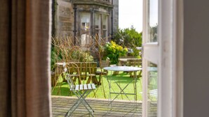 A view through a glass door of a courtyard with a table and chairs, a stone building in the background, and a grassy area.