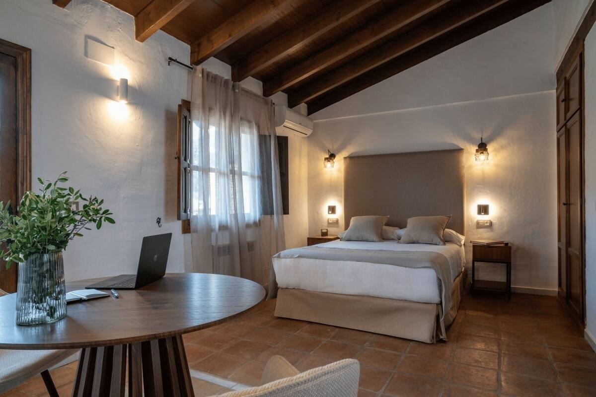 Une chambre à coucher avec un lit, des tables de chevet, un ordinateur portable posé sur une table ronde et une plante en pot.