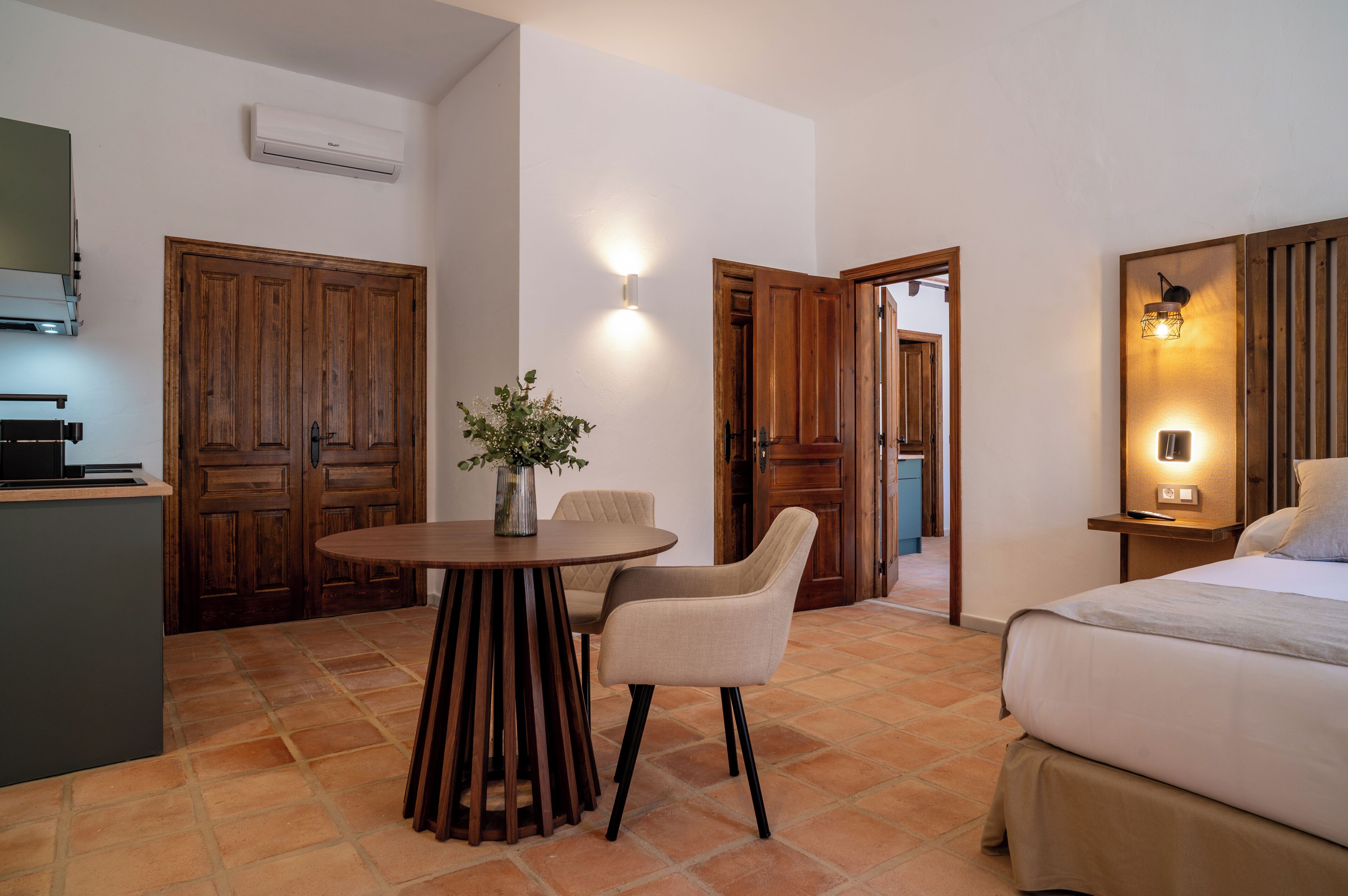 Habitación de hotel con cama, mesa de comedor con sillas, pequeña cocina y baño visible a través de una puerta abierta.