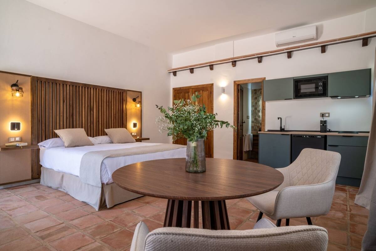 Une chambre avec un lit, des tables de chevet, une table ronde en bois et une chaise. Il y a également un petit coin cuisine équipé d’un four à micro-ondes et d’un évier.