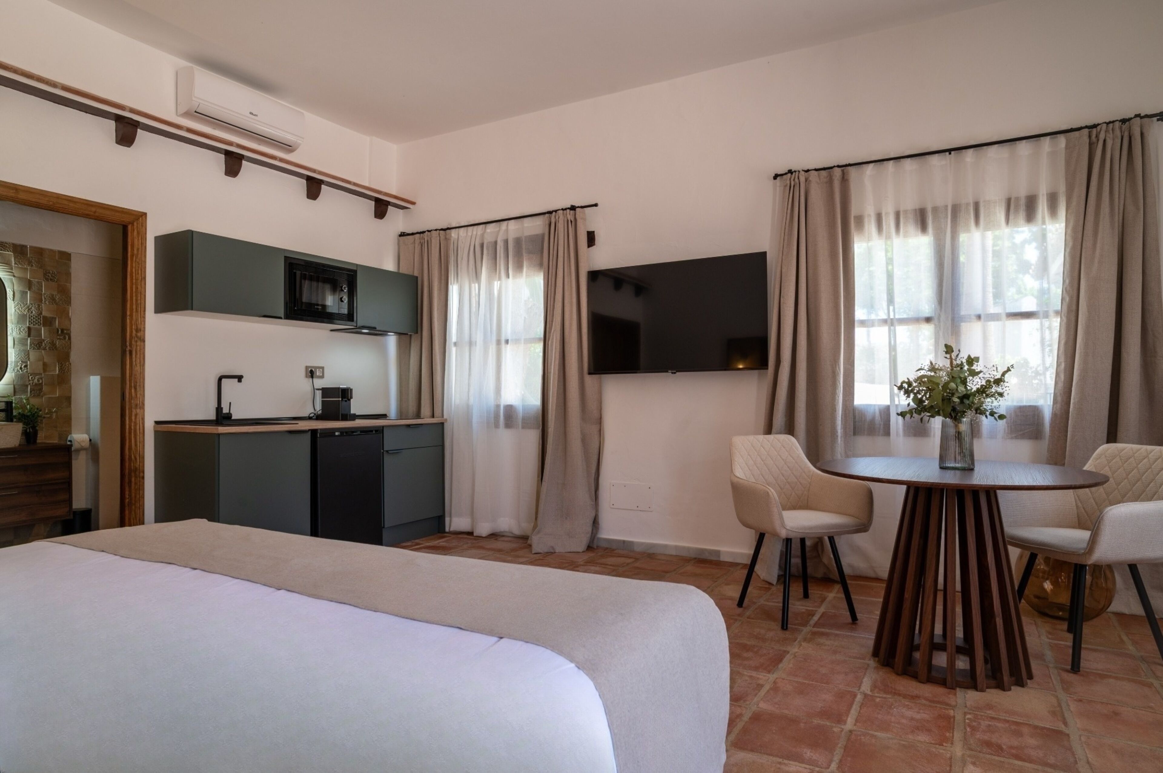 Une chambre d’hôtel comprenant un lit, une table à manger avec des chaises, un coin cuisine avec un four à micro-ondes et un téléviseur fixé au mur.