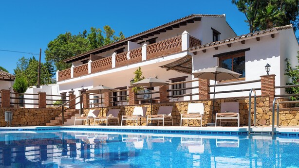 Zona de piscina con sillones de descanso y sombrillas, una pared de piedra y un edificio con tejado de azulejos y balcones.