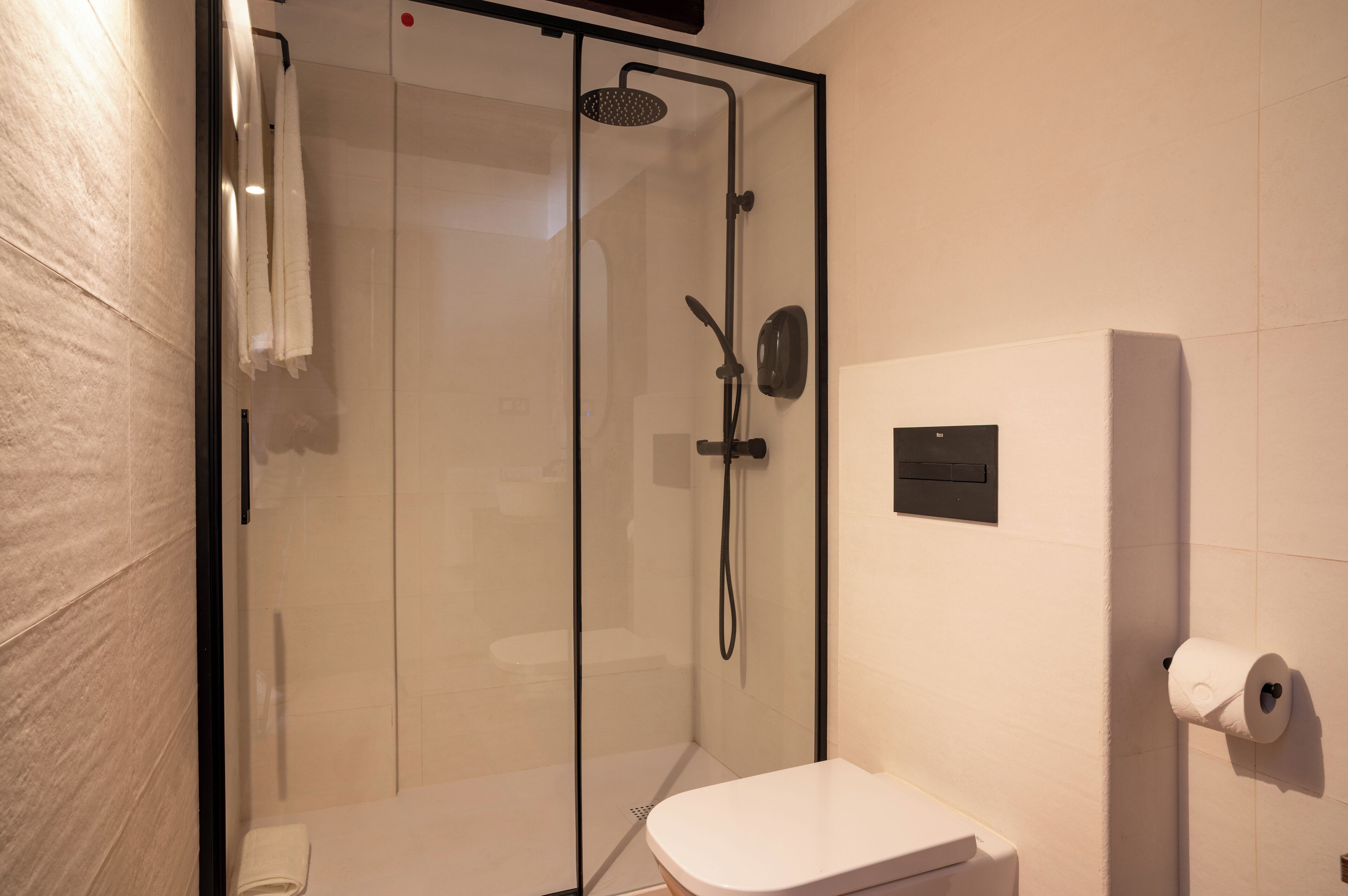 Un baño moderno con ducha de vidrio, inodoro y dispensador de papel higiénico en la pared.
