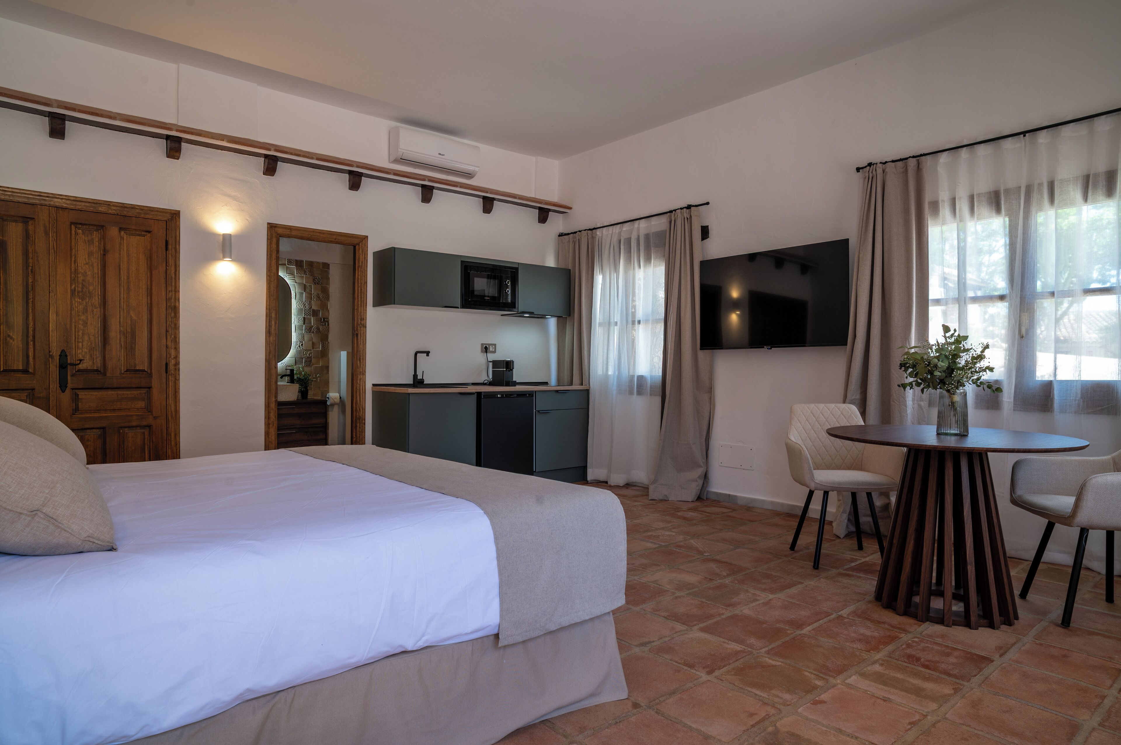A bedroom with a bed, a small kitchenette, a dining table with chairs, and a TV mounted on the wall.