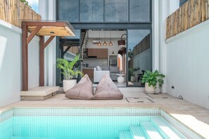 A modern outdoor pool area with a wooden pergola, seating cushions, and potted plants. - Villa Mogu (Canggu)
