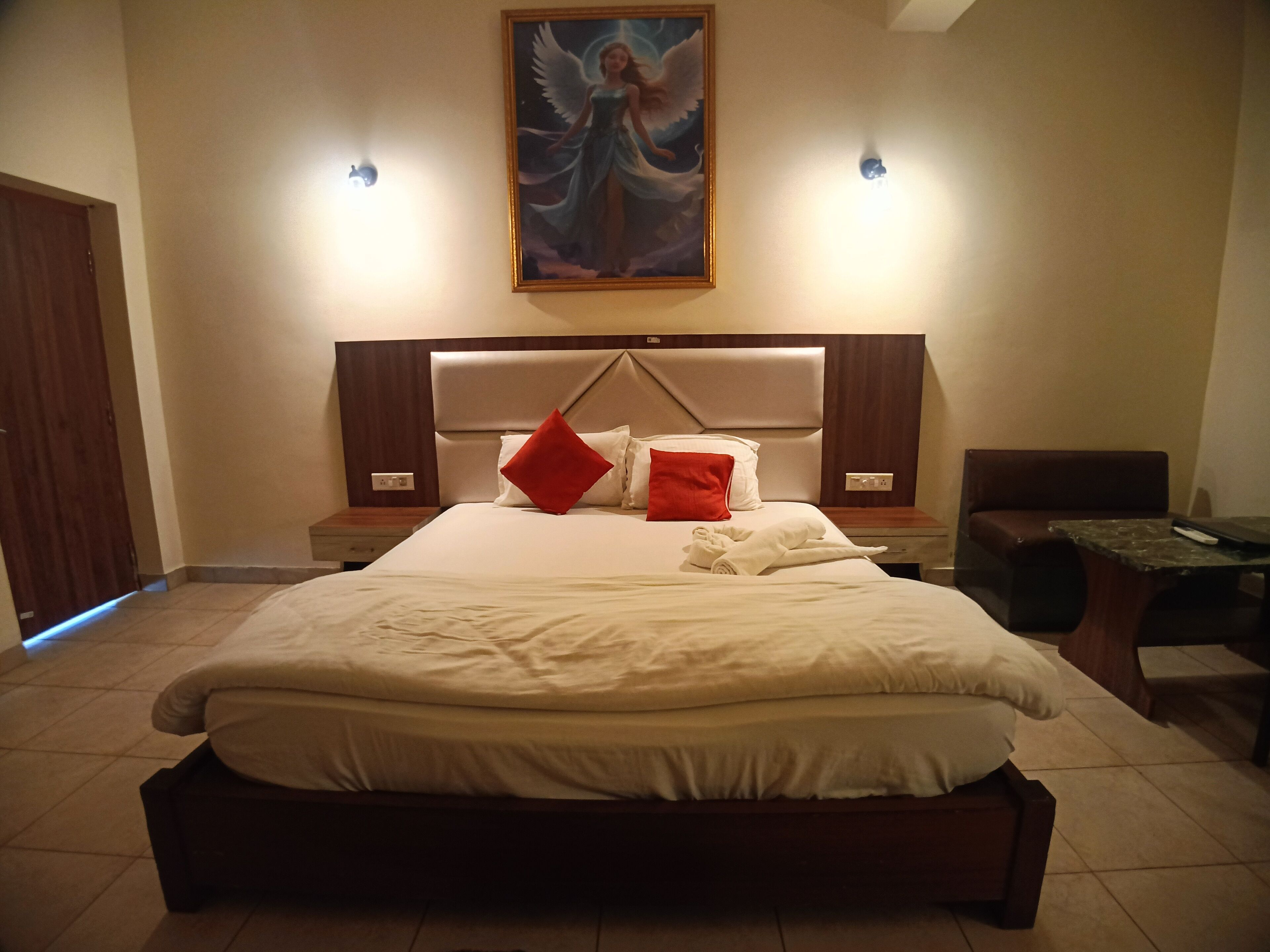 A neatly made bed with white linens and red pillows, a wooden headboard, a painting of an angel on the wall, and a small table with a remote control.