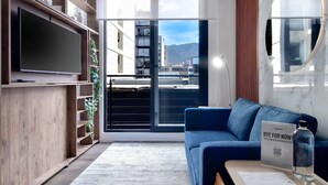 A modern living room with a TV, a blue sofa, a coffee table, and a view of buildings and mountains through a glass door. - W*| Amazing 1BR w/ Beautiful view in Virrey (Bogotá)