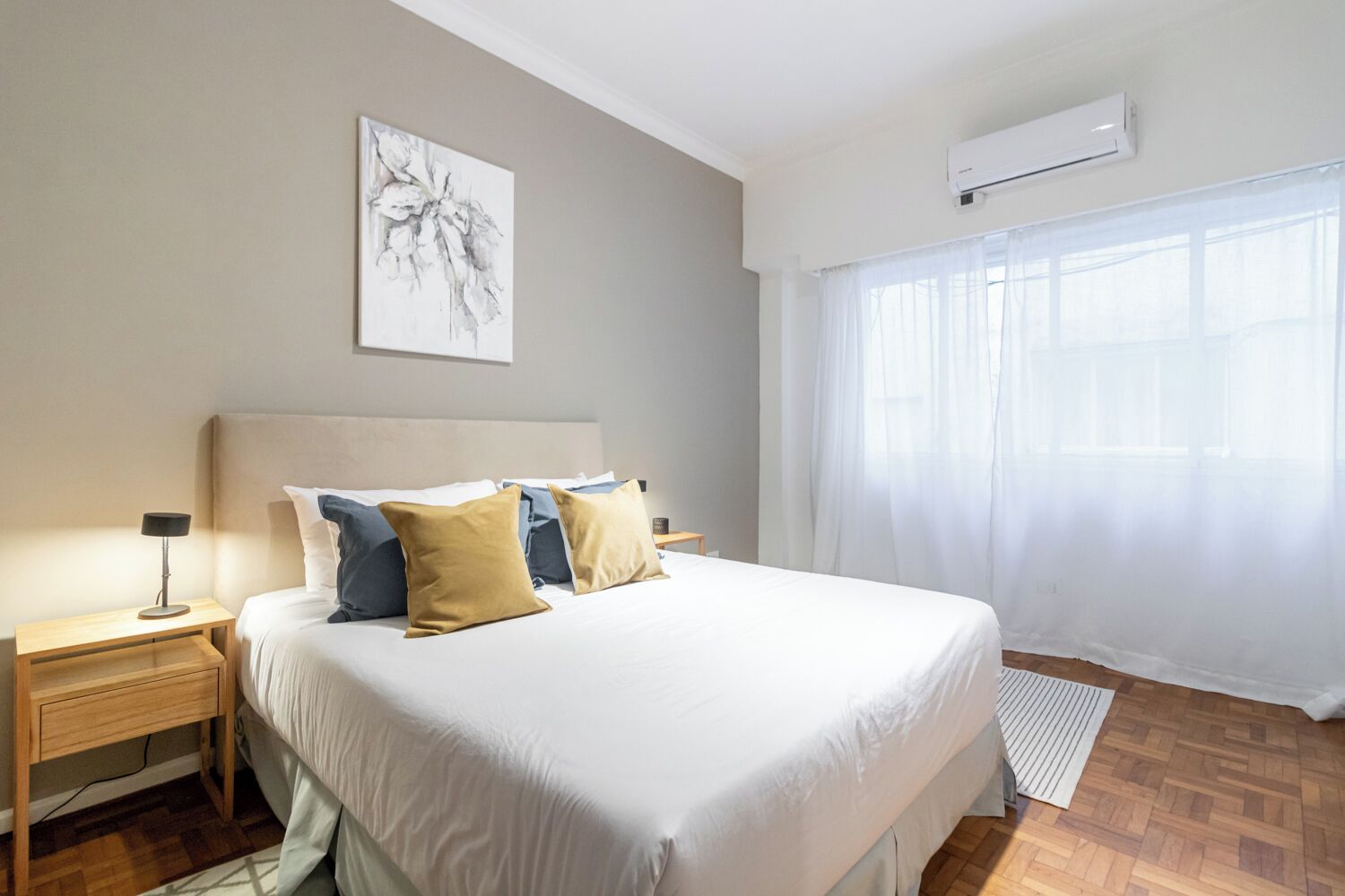Ein Schlafzimmer mit einem großen Bett, einem Nachttisch, einer Wand-Klimaanlage und einem Fenster mit durchsichtigen Vorhängen.