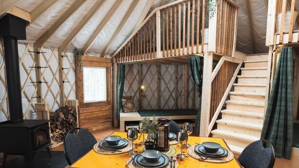 A cozy interior with a wooden table set for dining, a staircase, and a small seating area. - Cozy Yurt in the Woods (Sainte-Agathe-des-Monts)