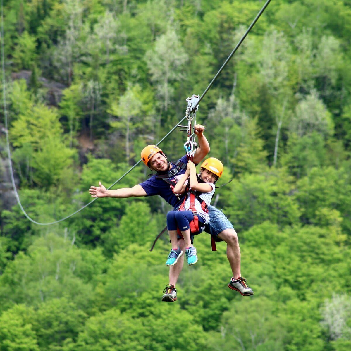 Zwei Personen mit Sicherheitsausrüstung beim Ziplining inmitten eines bewaldeten Gebiets.