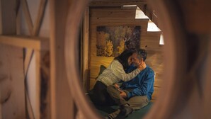 Pariskunta istuu puisen mökin sisällä olevalla penkillä, ja seinällä on taulu, jossa on kuvattuna puita.